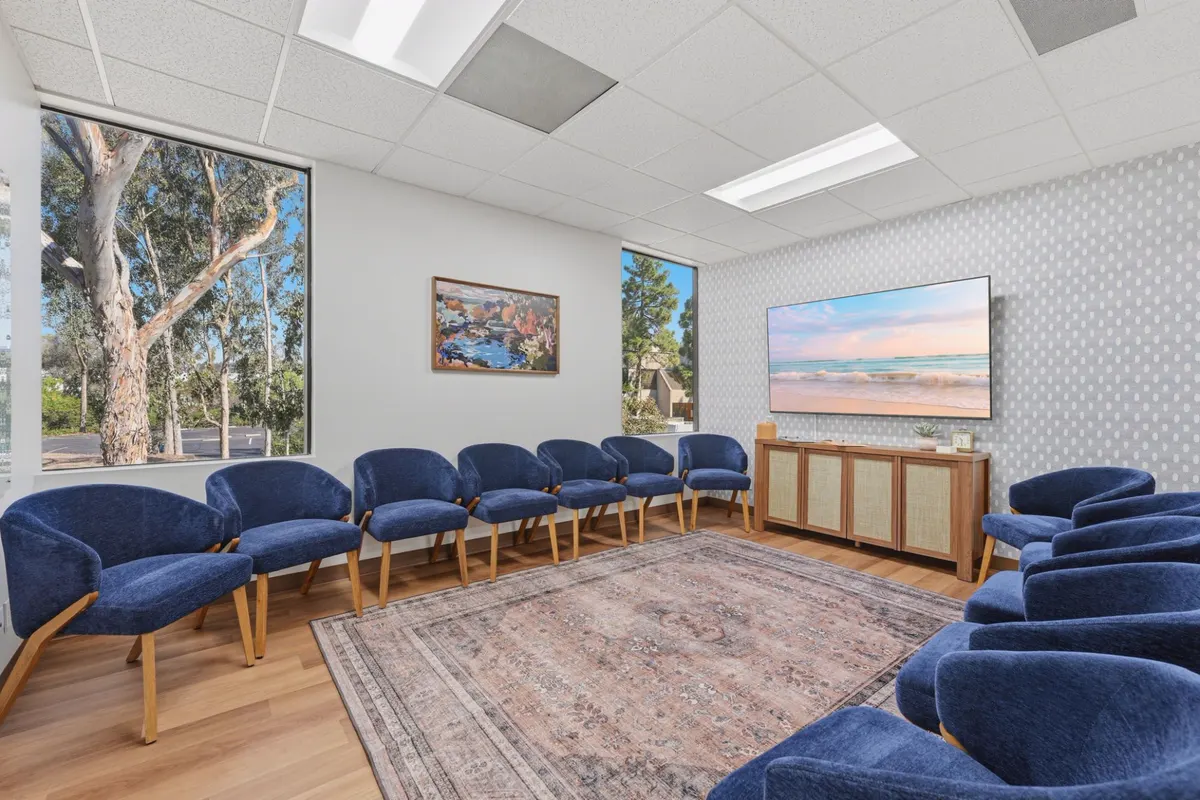 A group room with blue armchairs arranged in a circle around a patterned rug and windows framing a eucalyptus tree