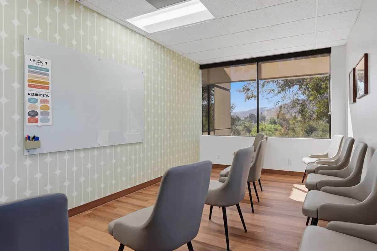 A group room with chairs in a circle and a glass whiteboard carrying check-in prompts on a patterned sage wall