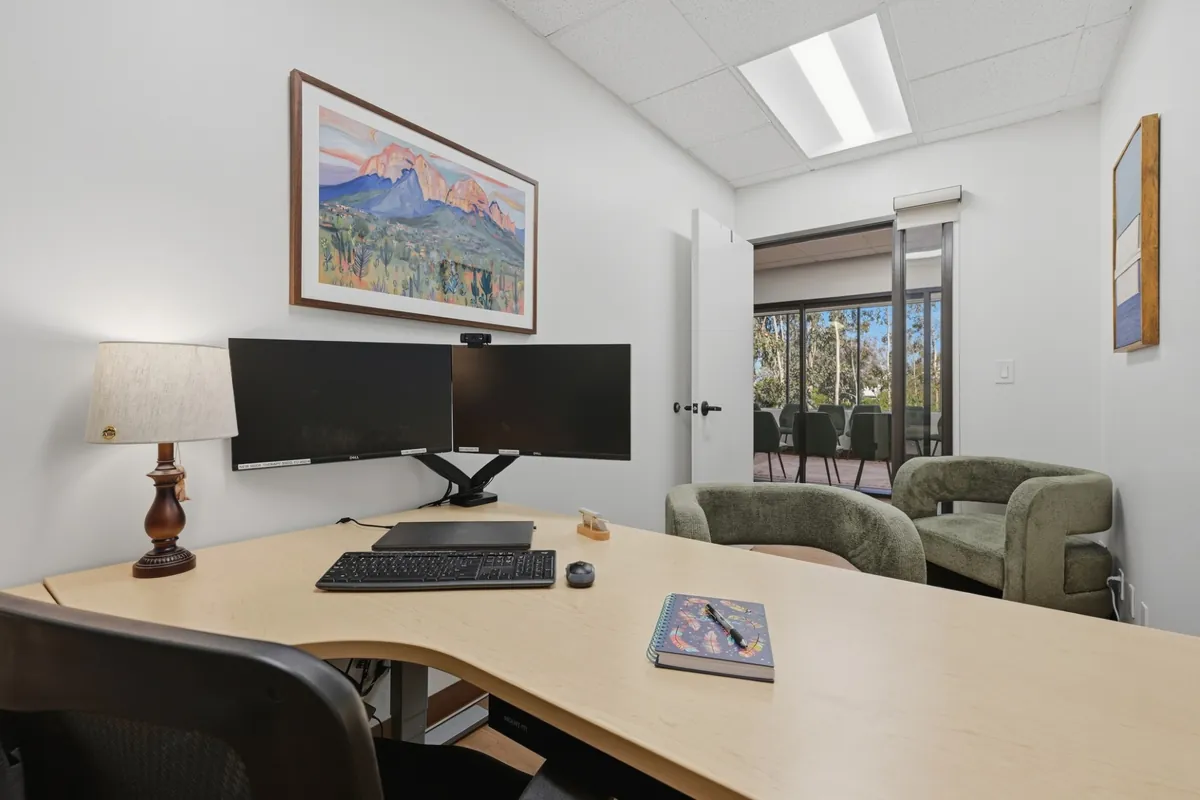 A clinician office with twin monitors and two green armchairs, its door open to a group room beyond