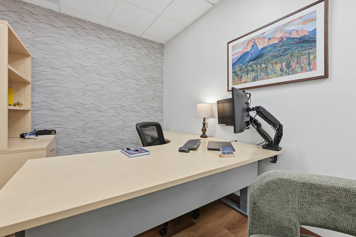 A clinician office with a pale wood desk, a monitor on an arm and a framed mountain print