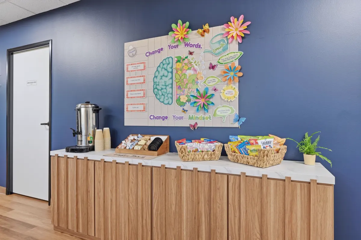 A refreshment counter with a coffee urn, cups, tea and snack baskets below a noticeboard on a deep blue wall